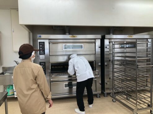 オーブンにパンを入れる作業をする利用者さんとそれを見守る職員の写真