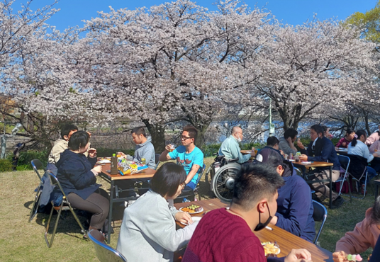 満開の桜の下でテーブルと椅子を広げてお菓子やジュースの飲むB型利用者さんたちの写真①
