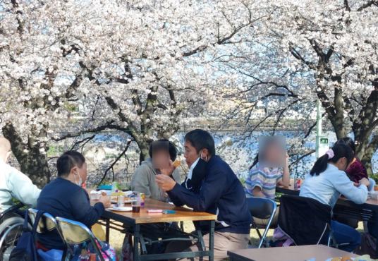 満開の桜の下でテーブルと椅子を広げてお菓子やジュースの飲むB型利用者さんたちの写真②